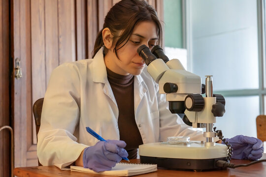 Female scientist looking through microscope and taking notes close-up