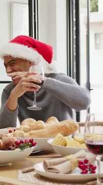 Vertical video: Laughing man in Santa hat holding wine glass reacting to offscreen joke at table