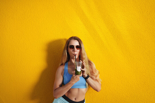 Happy woman with curly hair wearing sunglasses and holding a matcha cofe or milkshake against a yellow background.
