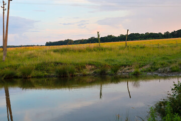 A small calm river flows past green pastures and spacious fields under the evening summer sky. The water surface softly reflects the distant forest line, wooden poles, and gentle shades of sunset clou © Олег Струс