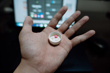 Fototapeta premium Hand holding small white pill cap with red tablet inside, blurred laptop background — close-up