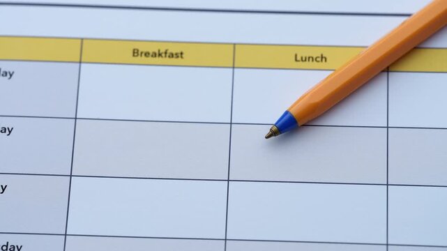 A blank weekly meal planner template with columns for breakfast, lunch, and dinner, featuring a yellow ballpoint pen resting on the grid. Close up.