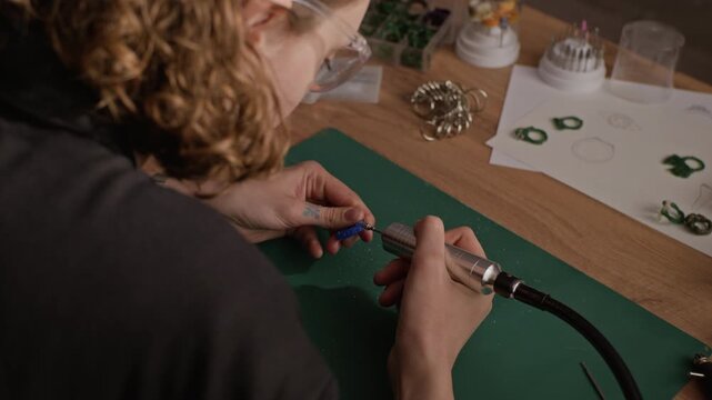 Over shoulder shot of Caucasian young adult female jeweler carving blue wax ring mold using rotary tool at workshop desk. Clip for promoting handmade jewelry brands, craft workshops, or design schools
