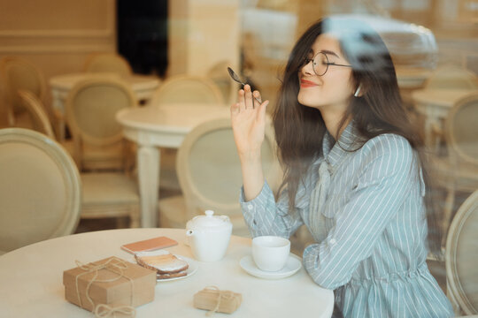 beautiful asian girl enjoying hot tea in cafe while listening music on headphones, view through window glass