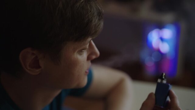 Close-up view of young adult man inhaling vapor from electronic cigarette, relaxing in dimly lit room with glowing computer case in background, modern smoking habit concept. Shooting in slow motion