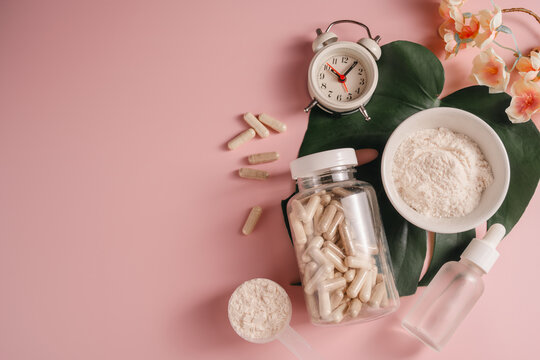 Collagen powder in a bowl with food supplement capsules, on monstera leaf with alarm clock on pink background, Healthy aging concept