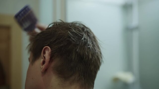 Close-up rear view of young man receiving haircut at home, with hand using comb and clipper to trim brown hair short. Concept of diy grooming process in bathroom setting from back view perspective