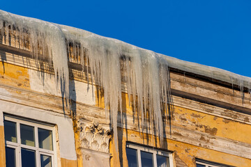Huge sharp icicles hanging from the crumbling yellow cornice of a historic building under a brilliant blue winter sky, showcasing frozen danger and cold weather © PhotoChur