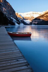 Fototapeta premium Tranquil lake reflects snow-capped mountains. Small red boat rests near wooden pier. Calm water mirrors peaks, creating stunning scenery. Early morning light illuminates landscape. Peaceful scene