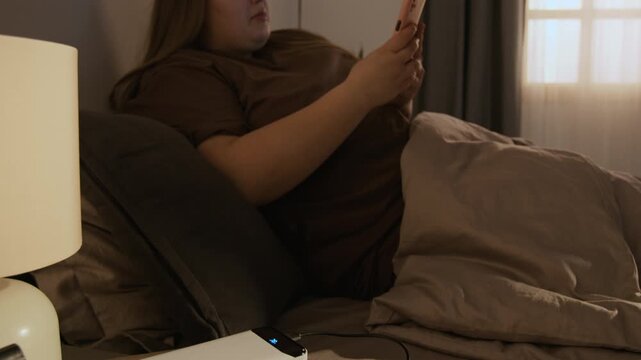 Plus-size Caucasian young adult woman unplugging smartphone from power bank and scrolling while lying in bed. Clip is suitable for mobile technology and morning routine commercials