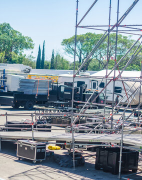 Outdoor concert stage setup backstage area with scaffolding flight cases trucks and crew vehicles preparing for a live event on a sunny day