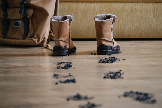 Mud-covered winter boots leaving dirty footprints on wooden floor indoors in living room. A backpack is seen in the background. Home care and cleaning concept.