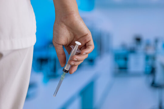 Hand grips a syringe. The syringe has a needle and plunger. Blurred background shows a medical lab setting. White scrubs are visible on the arm. This image captures healthcare professional work