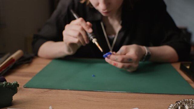 Close up of young adult Caucasian female jeweler carving blue wax ring mold using soldering tool at workshop table with sketches. Video for marketing handmade jewelry brands or craft workshops
