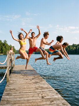 A group of friends leaps from a wooden dock into a peaceful lake, celebrating a carefree summer day together.