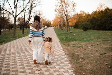 Fototapeta premium Happy mother with kid girl daughter walking in autumn park wearing knitted clothes over yellow leaves rear view. Fall season. Motherhood.