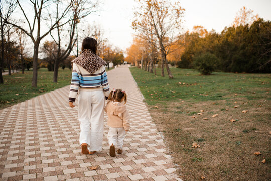 Happy mother with kid girl daughter walking in autumn park wearing knitted clothes over yellow leaves rear view. Fall season. Motherhood.