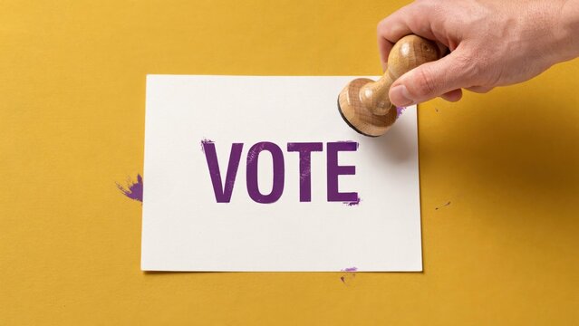 A hand stamps the word VOTE on a white paper with a wooden stamper on yellow background.