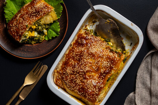 Homemade golden puff pastry pie filled with spinach, eggs and cheese, topped with sesame seeds and served fresh from baking dish, closeup, top view. Perfect for brunch, lunch or family dinner
