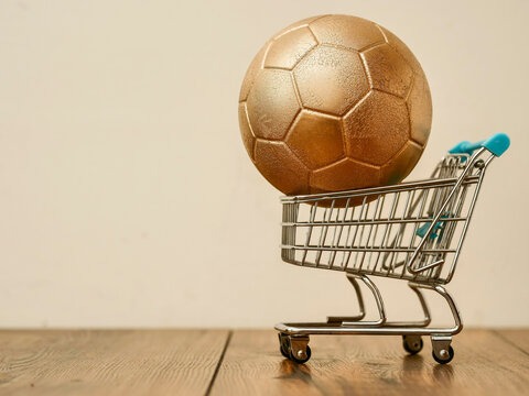 Golden soccer ball sits in tiny shopping cart. Cart rests on wooden surface. Simple background. Minimalist composition. Suitable for sports, commerce, or lifestyle themes