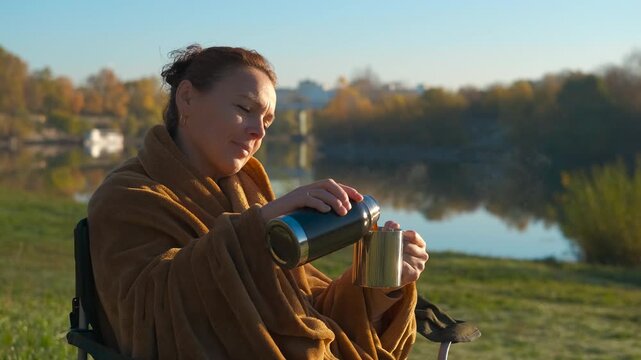 Woman pouring hot tea from thermos during autumn camping. Peaceful woman wrapped in a warm blanket pouring a steaming hot drink from a thermos into a cup while sitting on a camping chair by a river
