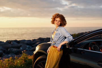 Fototapeta premium A stylish woman stands beside a parked car at golden hour by the sea, wearing a denim jacket and yellow dress. A lifestyle portrait conveying confidence and outdoor freedom.