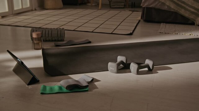 Fitness equipment lying on floor including yoga mat, dumbbells, resistance bands, and tablet during home workout, panning shot. Ideal for promoting online training apps and healthy lifestyle
