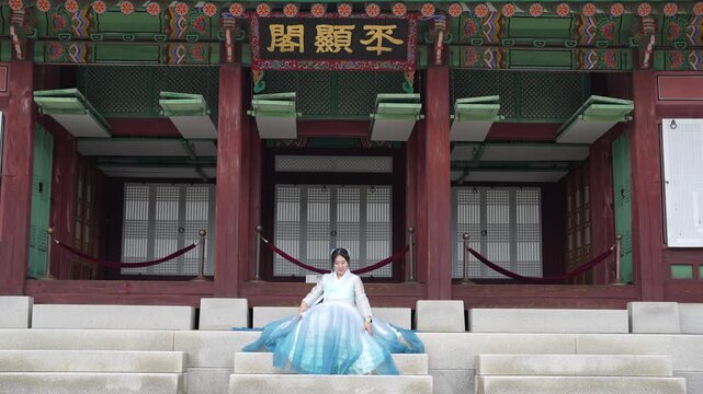Elegant woman in flowing blue hanbok sits gracefully. She extends arms outward, appearing serene and poised. Traditional Korean architecture frames her with calm dignity