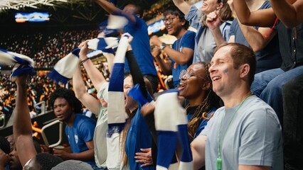Diverse blue team women supporters cheering loudly with raised arms in packed stadium stands during exciting live soccer match, Team spirit and celebration inside the stadium terrace. © DC Studio
