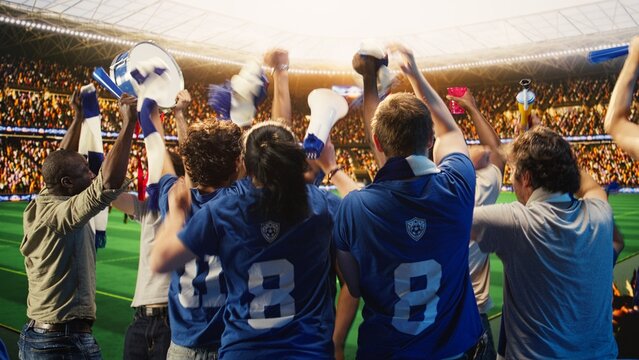 Rear view of happy football supporters in blue team jerseys raising fists to celebrate during soccer match day in packed stadium, soccer crowd jumping from joy after after competition victory.