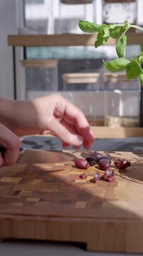 Hand slices olives on wooden cutting board, with basil leaves in the background. Concept of fresh ingredients and Mediterranean cooking