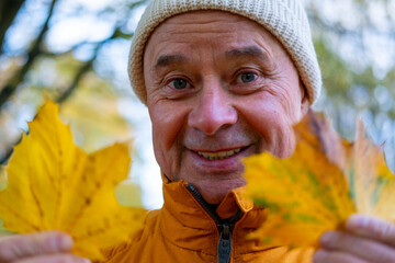 Smiling senior man in orange jacket holds bright yellow autumn leaves, looking joyfully at camera,...