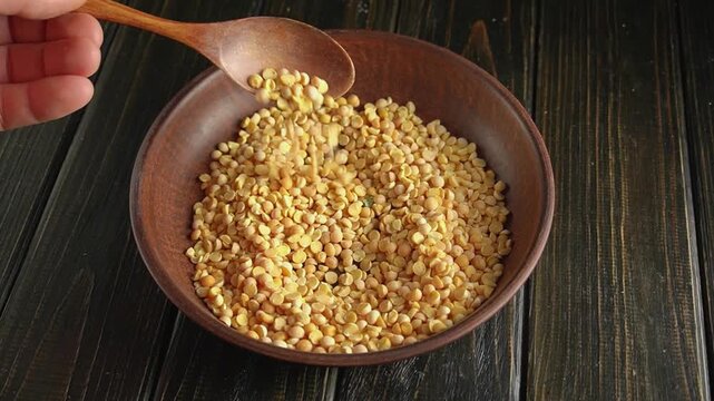 Hand scooping yellow split peas from a wooden bowl with a wooden spoon, showcasing the texture and color of the legumes on a dark wooden surface