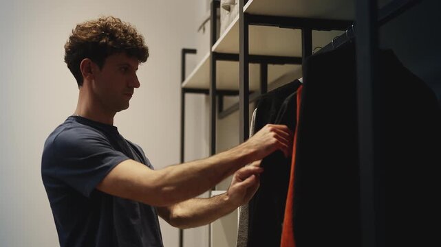 Curly-haired young man choosing between outfits beside wardrobe rack
