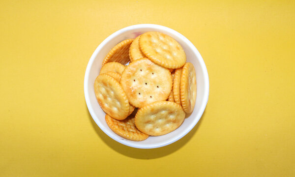 Stack of round salty crackers in a white bowl isolated on a yellow background. Top view Crispy baked biscuit snack with golden texture