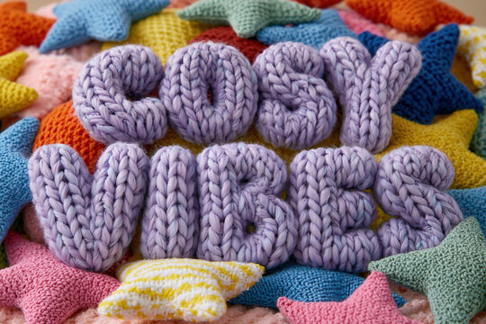 Cozy Knitted Serenity, the soft, lilac-colored text phrase "COSY VIBES" is beautifully knitted into thick woolen letters, resting comfortably on a pile of vibrant, multi-colored crochet stars