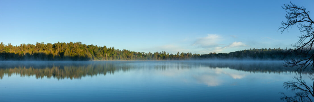 Panorama of a blue northern Minnesota lake and pines with morning fog under blue sky