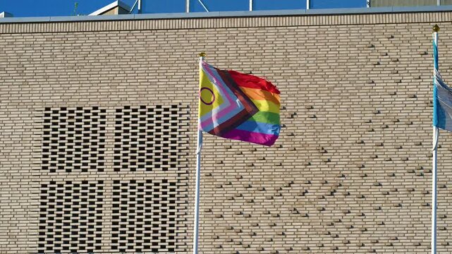 Progress Pride flag waving beside modern brick building in sunlight