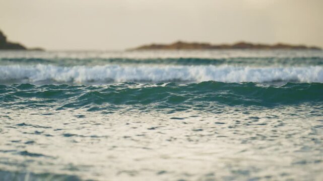 Rolling blue ocean wave before blurred island coastline in warm light