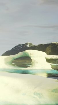 A large ice formation sits next to a calm body of water. Mountains surround the area. It is daytime with clear light shining on the ice structure and water.