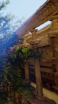 An old structure with tall, carved pillars. Green plants grow among the stones. The sunlight shines on the walls. People may explore this historic site.