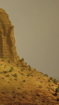 A large rock formation rises above the sandy landscape. Small shrubs grow in the dry earth. The scene shows an arid environment under a light sky.