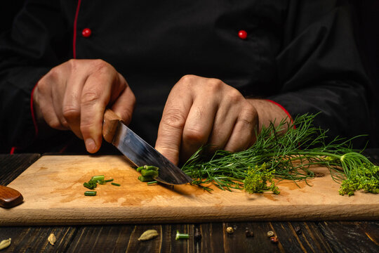 A chef uses a knife to chop fresh herbs dill on a wooden board. The chef's hands show focus and skill as they prepare ingredients for a meal. The kitchen has a warm atmosphere
