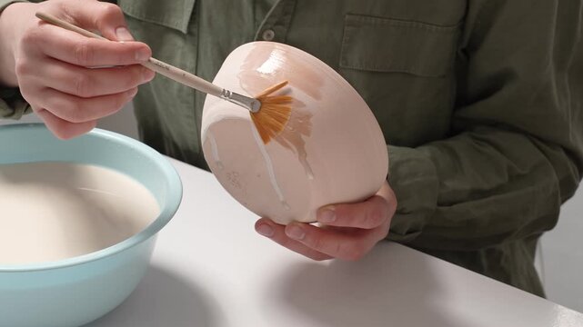Artisan Glazing Ceramic Bowl with a Brush. 4K Video artist uses a wide studio paintbrush to apply thick glaze to a bisque ceramic bowl for an artistic finish