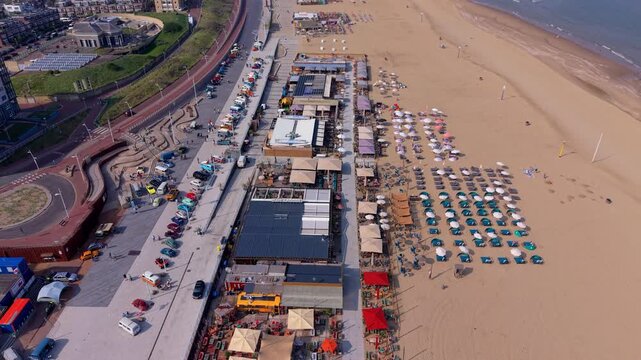 High-angle aerial view of a busy coastal beach resort featuring a long promenade with parked cars, vibrant beach club pavilions, and neat rows of umbrellas on the sand.