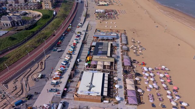 High-angle aerial view of a busy coastal beach resort featuring a long promenade with parked cars, vibrant beach club pavilions, and neat rows of umbrellas on the sand.