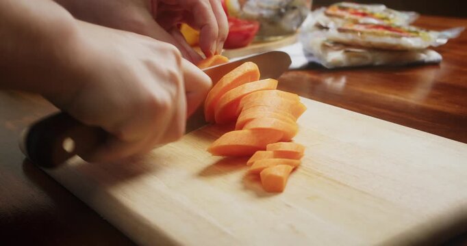 Close up of chef hands slicing fresh carrots on a wooden cutting board with a sharp kitchen knife. Fresh carrots are cut into diagonal pieces during food preparation in a home kitchen.