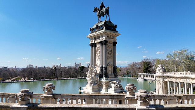 Wide aerial composition showing the rear side of the Monument to Alfonso XII overlooking Retiro Lake with Madrid skyline beneath a clear blue sky