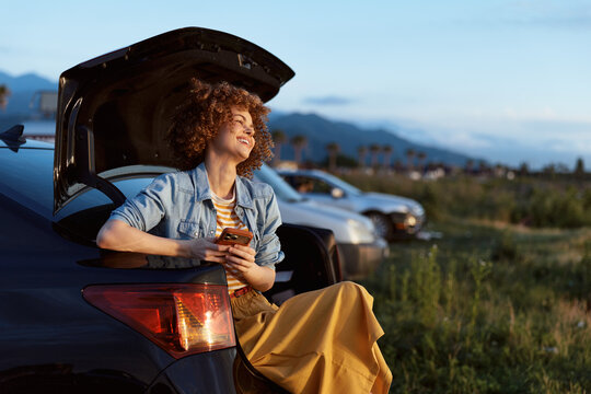 A smiling woman in a denim jacket sits on a car trunk at dusk, using a smartphone during a relaxed roadside moment, enjoying the calm landscape and casual travel vibe.