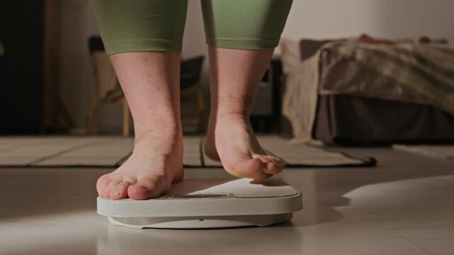 Plus-size Caucasian young adult woman stepping barefoot onto floor scale at home. Clip is perfect for weight loss programs, body positivity campaigns, and fitness tracking apps